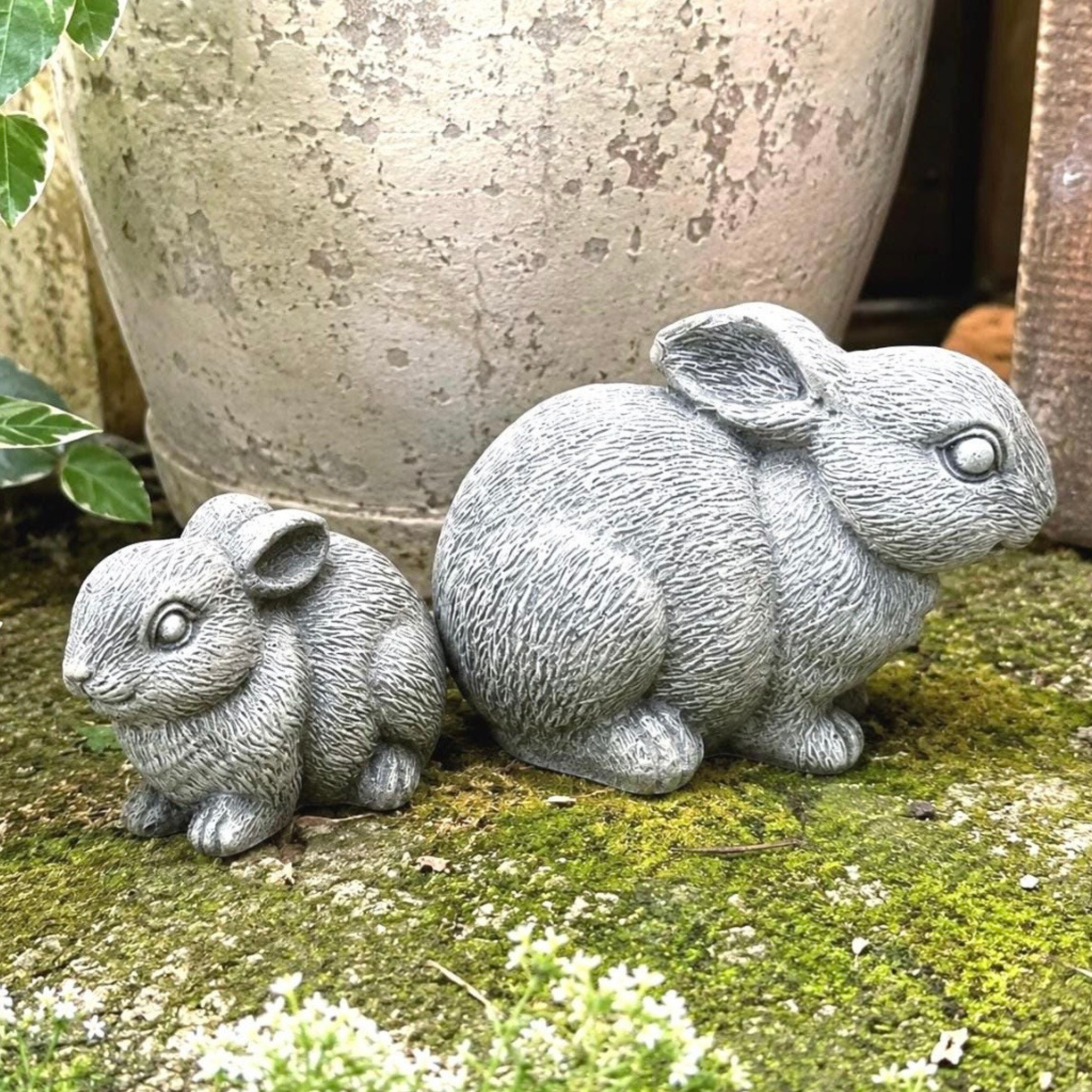 Two miniature stone rabbit sculptures on a mossy surface with a large pot in the background.