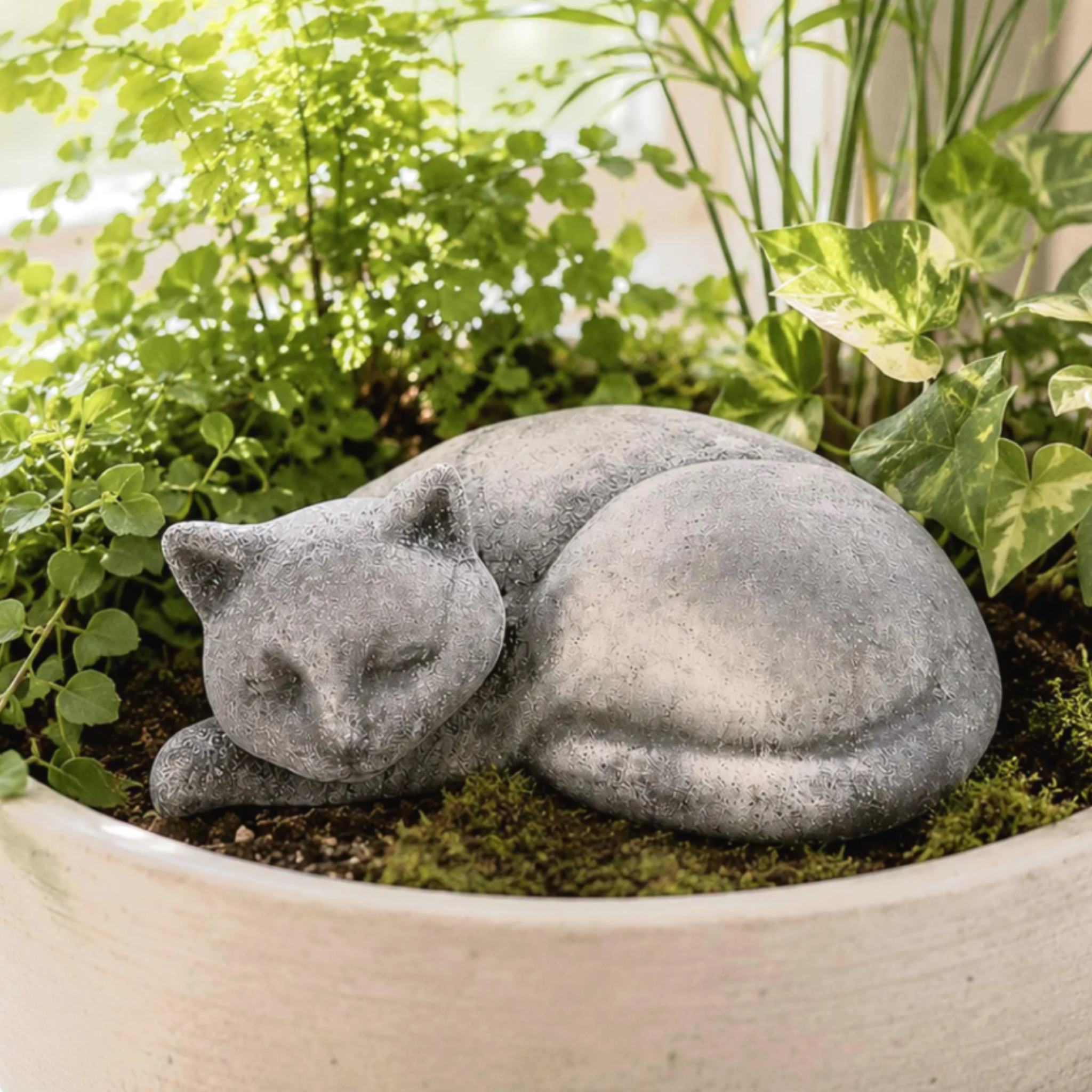 A gray, concrete statue of a sleeping cat with a smooth, rounded body and closed eyes. The cat figure is positioned resting on top of dark soil within a large, light-colored ceramic planter filled with various green plants.