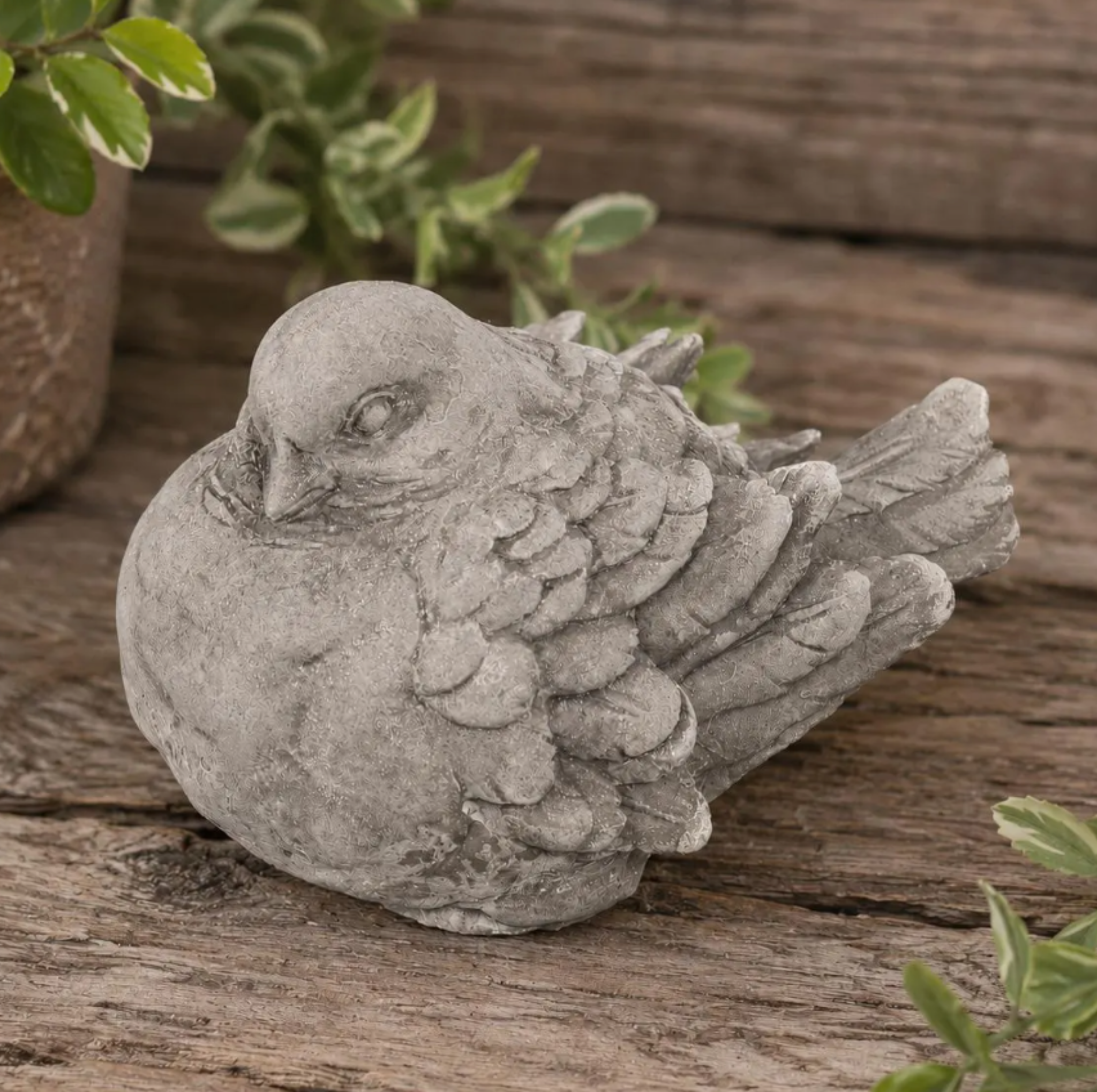 Stone concrete sculpture of a dove bird statue on a wooden surface with plants in the background