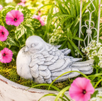 Whitewashed Outdoor Bird Dove Concrete Garden Statue perched inside a ceramic outdoor planter. The bird sculpture is surrounded by pink petunias and a wrought iron miniature fence is behind the statue. 